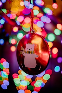 A festive ornament featuring a cartoonish polar bear with a striped scarf is hanging from a tree, surrounded by vibrant and colorful bokeh lights in the background.