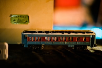 A model toy train carriage painted blue with red window frames and snow-like detailing on the roof. It is positioned on a black surface against a beige background with a slightly out-of-focus ticket marked 'Express' in the background. The scene is warmly lit, creating a cozy and nostalgic atmosphere.