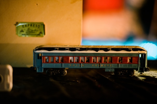 A model toy train carriage painted blue with red window frames and snow-like detailing on the roof. It is positioned on a black surface against a beige background with a slightly out-of-focus ticket marked 'Express' in the background. The scene is warmly lit, creating a cozy and nostalgic atmosphere.