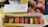 A selection of colorful macarons arranged in a cardboard box alongside a decorative box labeled 'Les incontournables de Paris' and an advertisement for Pierre Hermé's spring and summer collection. The macarons are displayed in a variety of vibrant colors.