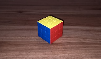 A partially solved 3x3 Rubik's Cube sits on a wooden surface. The visible sides show one face colored blue, another colored red, and the top face is yellow.