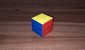 A partially solved 3x3 Rubik's Cube sits on a wooden surface. The visible sides show one face colored blue, another colored red, and the top face is yellow.