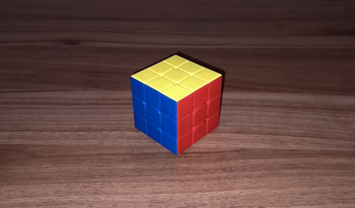 A partially solved 3x3 Rubik's Cube sits on a wooden surface. The visible sides show one face colored blue, another colored red, and the top face is yellow.