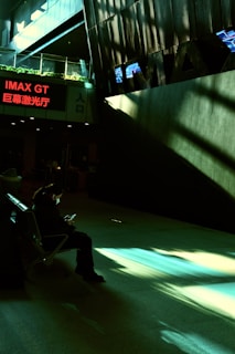 A person wearing a mask is seated in a dimly lit indoor area, possibly a cinema lobby, using a smartphone. Shadows and sunlight create patterns on the floor. The IMAX sign is visible with bright letters and a red neon sign displaying text in multiple languages.