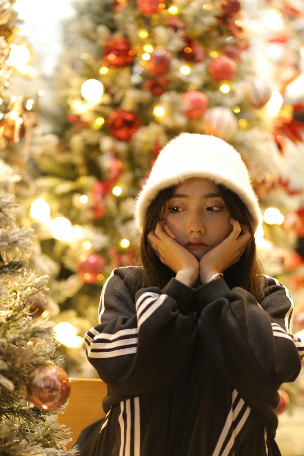 a young woman wearing a white hat standing in front of a christmas tree