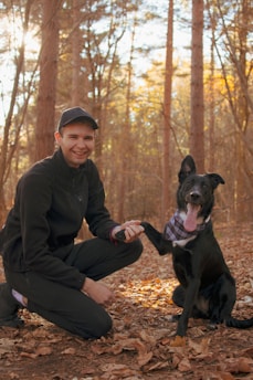 A person wearing a black jacket and cap is crouching down in a forest, holding the paw of a black dog wearing a plaid bandana. The ground is covered with autumn leaves and the sunlight filters through the trees, creating a warm glow.