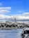 A coastal cityscape featuring a prominent historical bell tower rising above surrounding buildings. The waterfront is lined with palm trees and a row of boats docked in the harbor. The background showcases distant mountains under a partly cloudy sky with patches of blue.