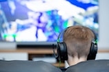 A person with short brown hair is sitting on a couch wearing black headphones. In front of them is a large screen displaying a video game with vibrant colors and dynamic visuals. The focus is on the person from behind, emphasizing the immersive gaming experience.