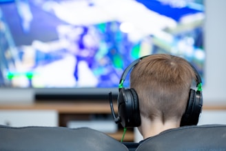 A person with short brown hair is sitting on a couch wearing black headphones. In front of them is a large screen displaying a video game with vibrant colors and dynamic visuals. The focus is on the person from behind, emphasizing the immersive gaming experience.