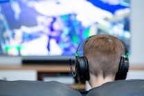 A person with short brown hair is sitting on a couch wearing black headphones. In front of them is a large screen displaying a video game with vibrant colors and dynamic visuals. The focus is on the person from behind, emphasizing the immersive gaming experience.