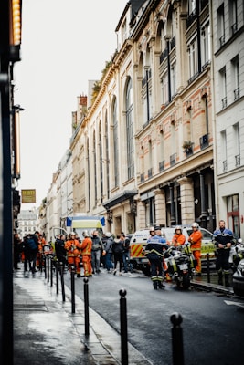 A city street with emergency responders gathered around. Several people in orange uniforms, possibly emergency medical technicians, interact with others on the scene. Police officers are present, as well as police vehicles and a motorcycle. The street is lined with tall, European-style buildings, and it appears to be a wet day, suggesting recent rain.