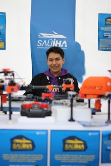 A young man is sitting and smiling at a booth with a blue backdrop that has the logo and name of Sakarya University. Various drones and related equipment are displayed on the table in front of him.