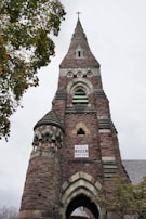 A tall church steeple made of stone rises into a cloudy sky, adorned with arched windows and ornate stonework. A 'Black Lives Matter' sign is prominently displayed on the front of the steeple. The structure is framed by leafy green branches on the left.