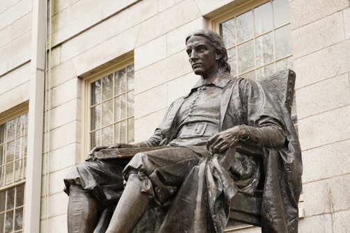 A bronze statue of a solemn figure seated in a chair against a brick building backdrop, with windows featuring grid-like panes. The figure wears historical attire with a detailed robe and holds a book or document in one hand.