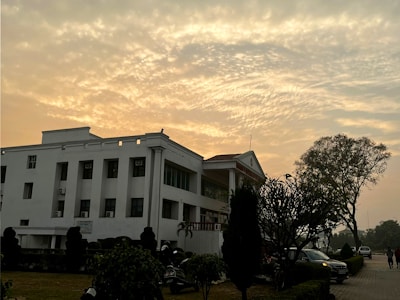 A white multi-story building with a flat roof stands in a landscaped area, featuring tall trees, shrubs, and a well-maintained path. The sky is filled with a dramatic pattern of clouds, illuminated by the warm glow of a setting or rising sun.