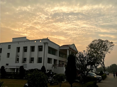 A white multi-story building with a flat roof stands in a landscaped area, featuring tall trees, shrubs, and a well-maintained path. The sky is filled with a dramatic pattern of clouds, illuminated by the warm glow of a setting or rising sun.