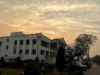 A white multi-story building with a flat roof stands in a landscaped area, featuring tall trees, shrubs, and a well-maintained path. The sky is filled with a dramatic pattern of clouds, illuminated by the warm glow of a setting or rising sun.