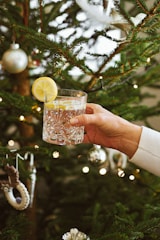 A hand holding a clear, intricately designed glass containing a beverage with lemon slices. The background features a decorated Christmas tree adorned with silver and white ornaments and fairy lights.