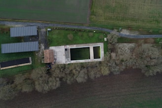 An aerial view of a rural area featuring rectangular buildings, solar panels, and paved pathways. The center shows a large concrete structure with two rectangular openings, surrounded by green grass and trees. Adjacent are two smaller buildings with a tiled roof, and a road running parallel to the structures.