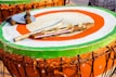 A large, round drum with a brightly painted surface featuring green, orange, and white concentric circles. Atop the drum are various percussion instruments, including sticks wrapped in fabric, a metal disc, and a wooden rod with cymbal-like attachments.