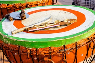 A large, round drum with a brightly painted surface featuring green, orange, and white concentric circles. Atop the drum are various percussion instruments, including sticks wrapped in fabric, a metal disc, and a wooden rod with cymbal-like attachments.
