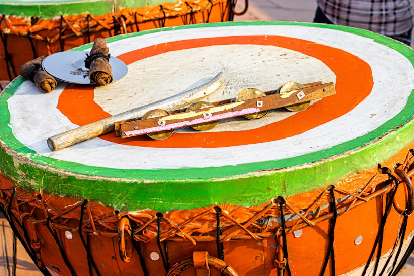 A large, round drum with a brightly painted surface featuring green, orange, and white concentric circles. Atop the drum are various percussion instruments, including sticks wrapped in fabric, a metal disc, and a wooden rod with cymbal-like attachments.