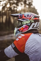 A motorcyclist wearing a colorful helmet with palm tree graphics, a patterned jersey predominantly in orange and white, in a forested area with blurred trees in the background.