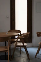 A minimalist interior setting featuring a small round wooden table surrounded by four wooden chairs with a modern design. The furniture is placed on a smooth gray floor, and a tall wooden-framed window in the background lets in soft natural light. A small glass candle holder is positioned on the table, adding to the serene atmosphere.