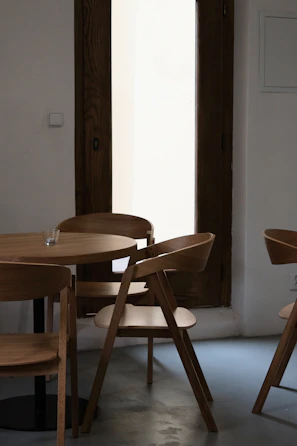A minimalist interior setting featuring a small round wooden table surrounded by four wooden chairs with a modern design. The furniture is placed on a smooth gray floor, and a tall wooden-framed window in the background lets in soft natural light. A small glass candle holder is positioned on the table, adding to the serene atmosphere.