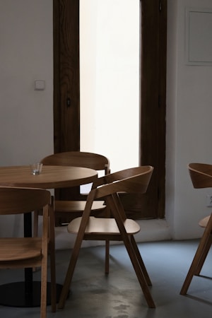 A minimalist interior setting featuring a small round wooden table surrounded by four wooden chairs with a modern design. The furniture is placed on a smooth gray floor, and a tall wooden-framed window in the background lets in soft natural light. A small glass candle holder is positioned on the table, adding to the serene atmosphere.
