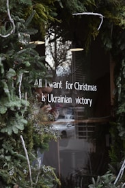 A window is decorated with green foliage and white text that reads 'All I want for Christmas is Ukrainian victory.' The background includes a reflection of the outside street and buildings.