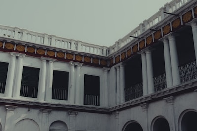 An old architectural structure with multiple levels, featuring columns and a series of windows and arches. The building has a classic design with ornate decorations, including yellow floral patterns above the columns.