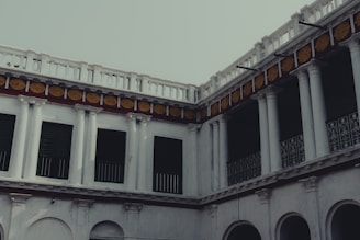 An old architectural structure with multiple levels, featuring columns and a series of windows and arches. The building has a classic design with ornate decorations, including yellow floral patterns above the columns.