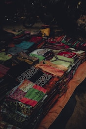 A collection of colorful folded fabrics displayed on a surface. The fabrics feature intricate patterns and a variety of colors, including green, red, black, and orange. They are arranged in neat piles, suggesting a market or shop setting.