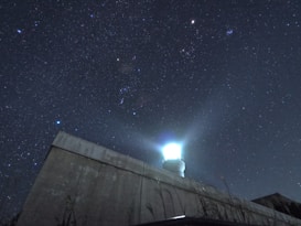 A starry night sky filled with numerous bright stars, with a prominent light source on top of a building or structure that appears to be a lighthouse or a similar installation. The light emits a strong glow against the dark sky, creating a striking contrast.