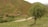 A picturesque rural landscape featuring a small village nestled among green hills. A winding road leads through the scene, bordered by vibrant trees and patches of grass. The earth-toned houses blend seamlessly with the natural surroundings, and the overall environment is serene and untouched.
