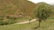 A picturesque rural landscape featuring a small village nestled among green hills. A winding road leads through the scene, bordered by vibrant trees and patches of grass. The earth-toned houses blend seamlessly with the natural surroundings, and the overall environment is serene and untouched.