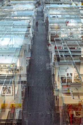 A complex, layered view of an interior space showcasing a modern art gallery or exhibition. Glass walls and partitions create a maze-like effect, with framed artworks visible on the walls. The perspective stretches into the distance, leading the eyes along a central path where a lone figure is walking.