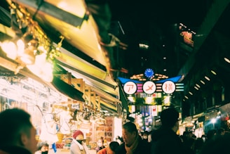 A bustling night market scene with bright lights and various people walking and shopping. The atmosphere is vibrant, with rows of stalls displaying goods and a prominent illuminated sign featuring Japanese characters.