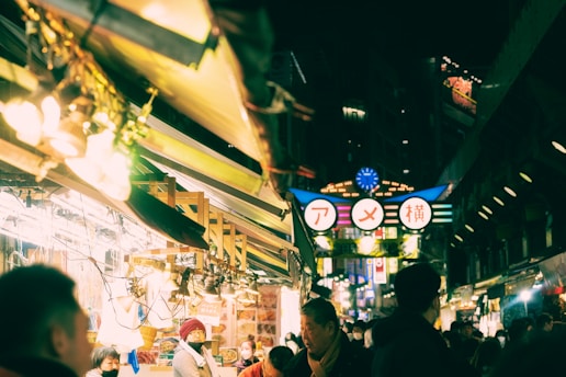 A bustling night market scene with bright lights and various people walking and shopping. The atmosphere is vibrant, with rows of stalls displaying goods and a prominent illuminated sign featuring Japanese characters.