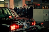A nighttime urban street scene with a black car and a small delivery vehicle. The car's taillights are illuminated, casting a red glow and reflecting off nearby surfaces. The delivery vehicle, with a large box or container attached to the back, is partially visible and driven by a person wearing dark clothing. In the background, there are lit storefronts and a crowd of people, with a warm ambient light creating a moody atmosphere.