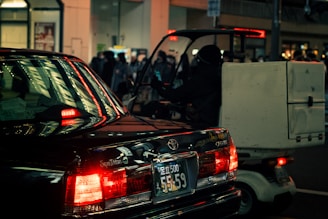 A nighttime urban street scene with a black car and a small delivery vehicle. The car's taillights are illuminated, casting a red glow and reflecting off nearby surfaces. The delivery vehicle, with a large box or container attached to the back, is partially visible and driven by a person wearing dark clothing. In the background, there are lit storefronts and a crowd of people, with a warm ambient light creating a moody atmosphere.