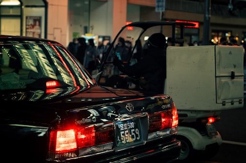A nighttime urban street scene with a black car and a small delivery vehicle. The car's taillights are illuminated, casting a red glow and reflecting off nearby surfaces. The delivery vehicle, with a large box or container attached to the back, is partially visible and driven by a person wearing dark clothing. In the background, there are lit storefronts and a crowd of people, with a warm ambient light creating a moody atmosphere.