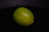 A single lime resting on a dark, possibly black, surface. The lime appears fresh with a smooth, glossy skin and a vibrant green color.