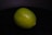 A single lime resting on a dark, possibly black, surface. The lime appears fresh with a smooth, glossy skin and a vibrant green color.