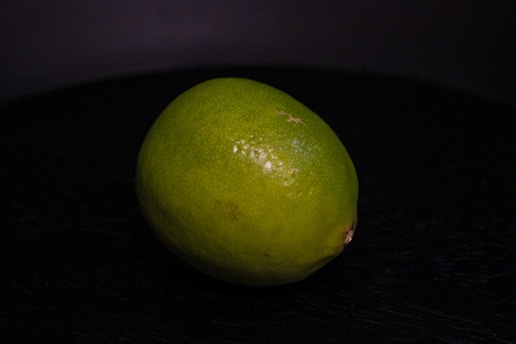 A single lime resting on a dark, possibly black, surface. The lime appears fresh with a smooth, glossy skin and a vibrant green color.