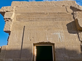An ancient stone wall features intricate carvings and hieroglyphs. The stone is weathered, displaying detailed depictions of figures and symbols characteristic of ancient Egyptian art. Bright blue sky contrasts with the warm tones of the stone.