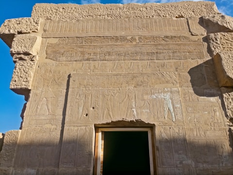 An ancient stone wall features intricate carvings and hieroglyphs. The stone is weathered, displaying detailed depictions of figures and symbols characteristic of ancient Egyptian art. Bright blue sky contrasts with the warm tones of the stone.