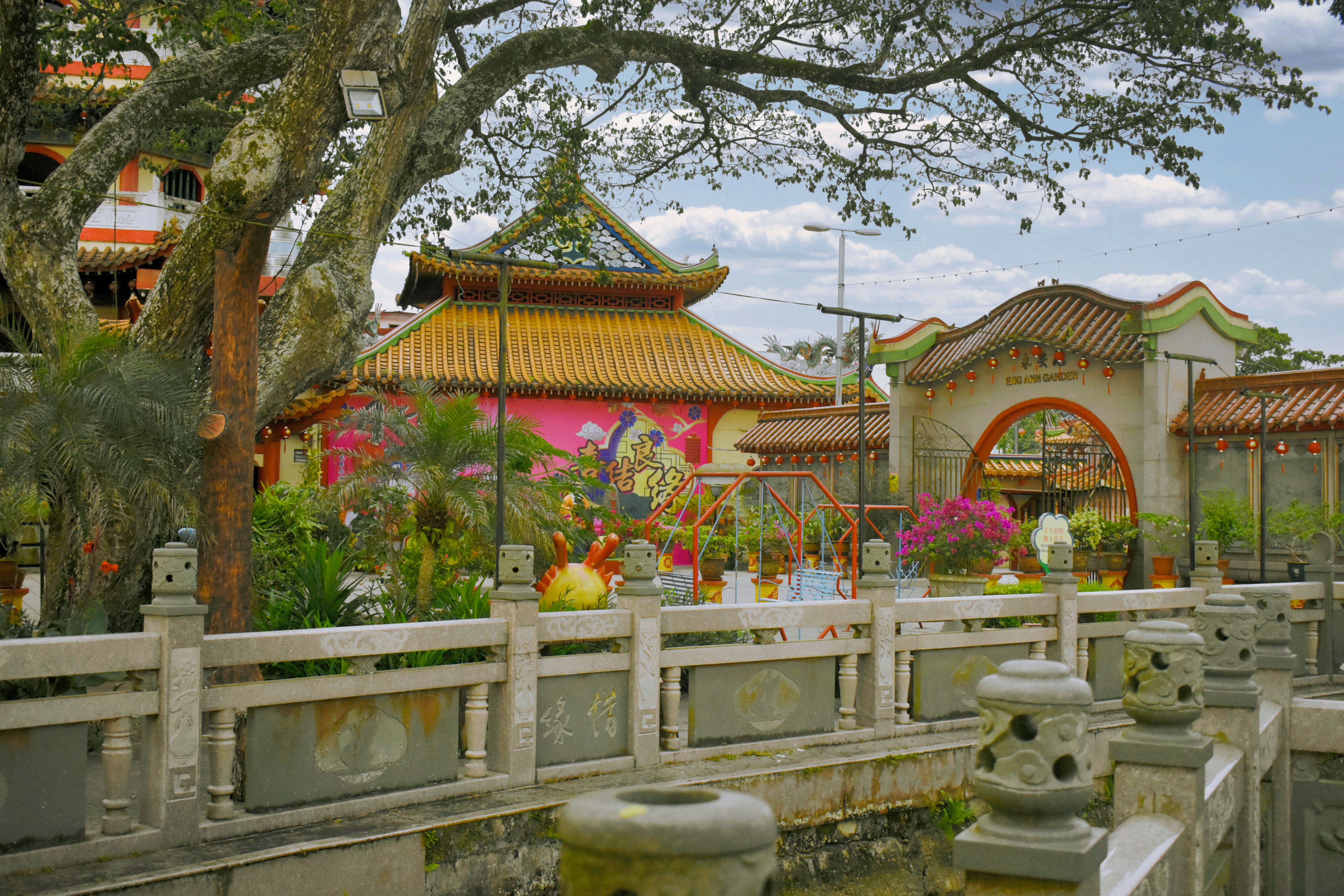 A tranquil temple courtyard with vibrant mural walls and ornate gates is framed by a stone railing along a canal. Blooming plants brighten the scene and highlight architectural detail.