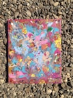 An abstract painting with vibrant swirling colors including blue, pink, orange, and yellow, placed on a background of small, smooth rocks.